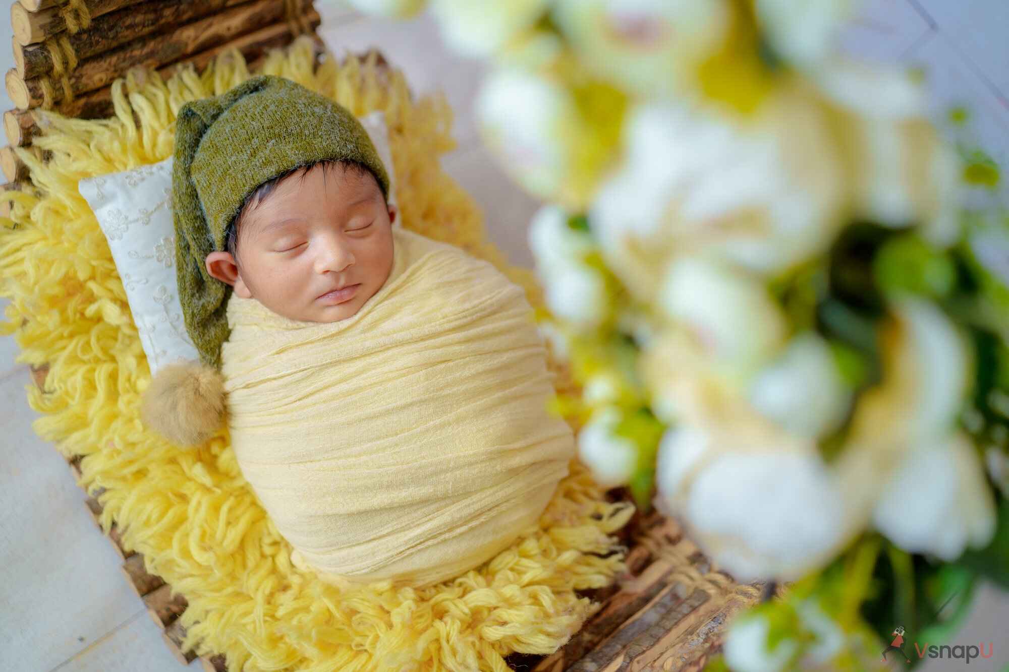 A baby napping in a wooden floral basket with a yellow fur mattress and surrounded by soft flowers in a lovely baby photoshoot.